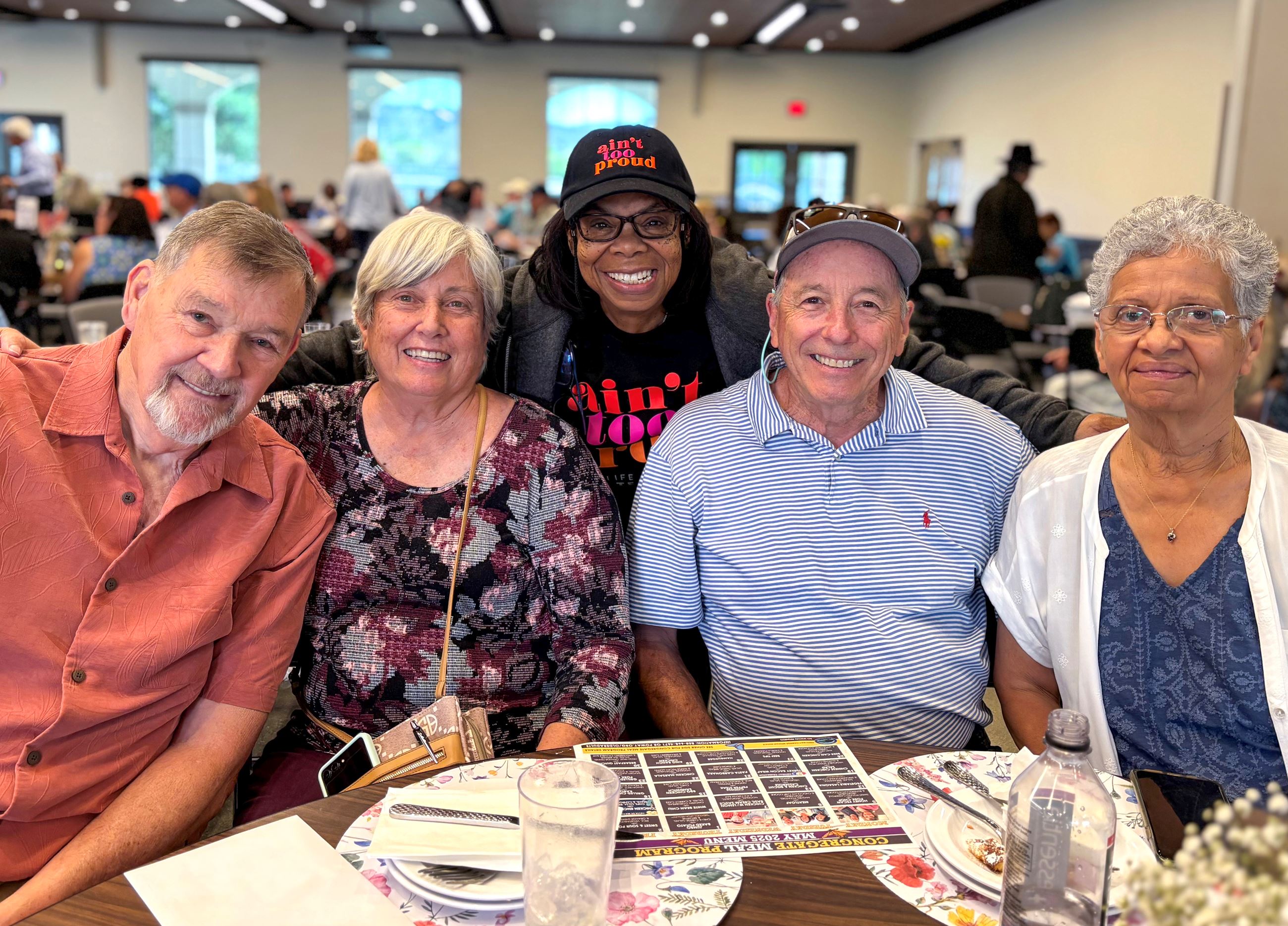 A group of two older men and three older women standing around a lunch table smiling